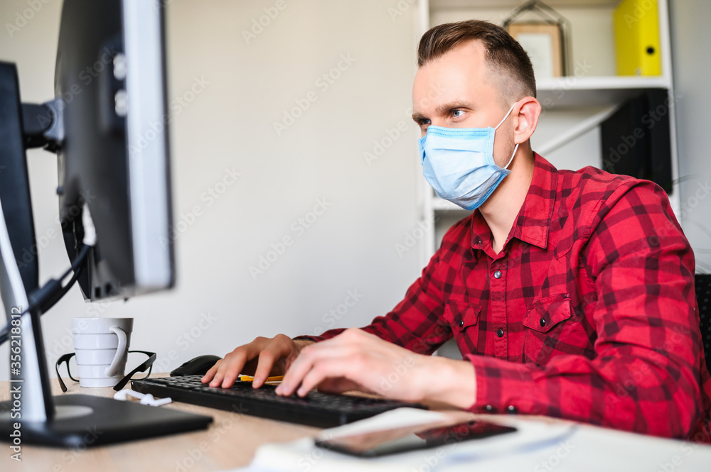 Responsible employee works with the computer wearing a protective mask. A young man works in the office while quarantine. Responsible employee works with the computer wearing a protective mask. A young man works in the office while quarantine.