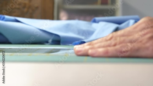 Side shot of female hand holding a ruler that guides a rotary cutter as it cuts blue cotton fabric on a green cutting mat. Pile of blue fabric laying in the background.