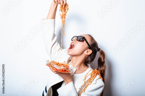 Sexy seductive provocative girl in clothes and a plate with pasta and a fork wildly eats pasta with ketchup  standing in glasses on a white background. Girl eating spaghetti.