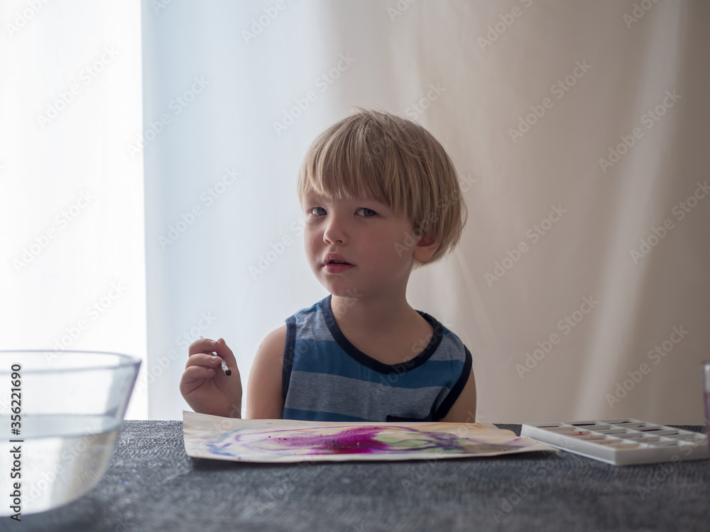 paintig lovely blond child with watercolors. Paints in cuvettes, brushes, water for wetting paper. boy likes to spend time at home. Wet paper produces beautiful patterns