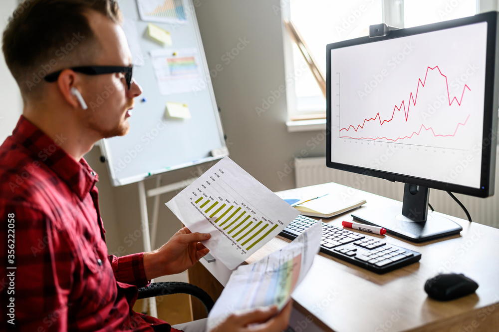 Concentrated guy lookolds printed s on the graph on PC monitor in front of him in the office and holds printed gistograms, he wears glasses and casual clothes, flipchart on background Concentrated guy lookolds printed s on the graph on PC monitor in front of him in the office and holds printed gistograms, he wears glasses and casual clothes, flipchart on background