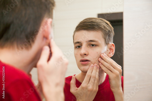 Close up of young attractive man with problematic skin and scars from acne looks in the mirror in the bathroom