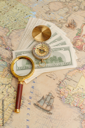 A bundle of American dollars lies with a compass and a magnifying glass on the world map