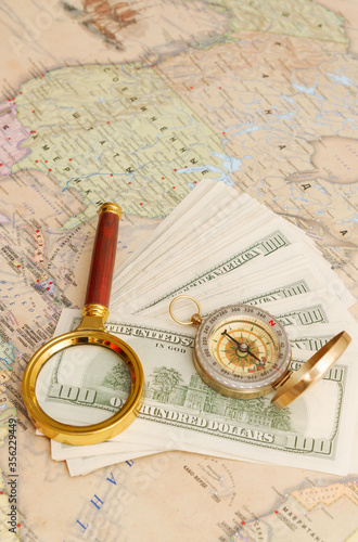 A bundle of American dollars lies with a compass and a magnifying glass on the world map