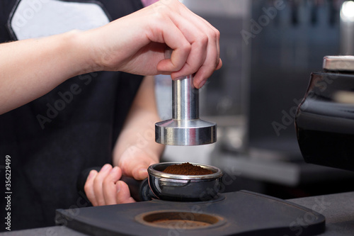 close-up photo of female hands holding a metal tamper and a portafilter with coffee in a coffee shop. A barista preparing for pressing ground coffee for brewing espresso or americano in a cafe