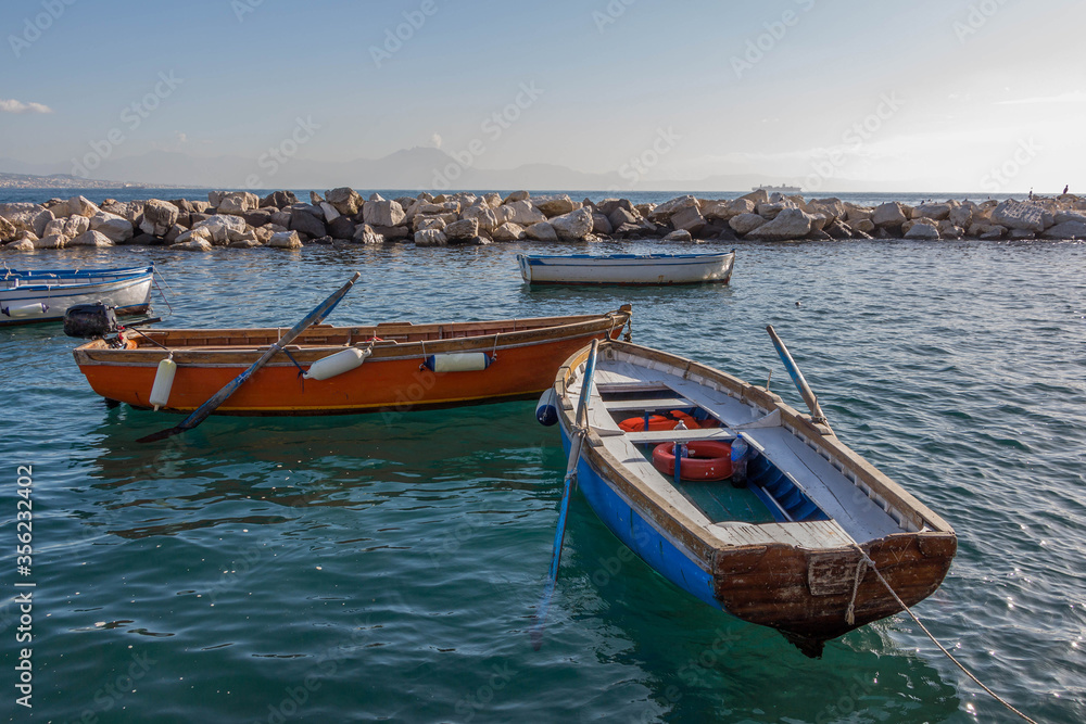 Fishing wooden boats with oars and motor in the turquoise water of the Bay of Naples against the background of a stone breakwater, in the haze of the mountains and the ship. Birds on the rocks.