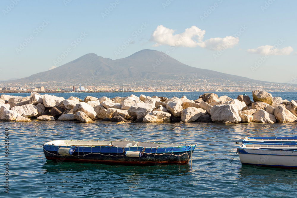 Boats in the turquoise Bay of Naples against the background of the stone coastline lit by the sun and the Vesuvius volcano with blue sky and white clouds