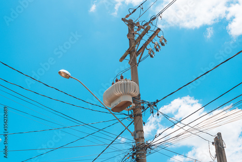 Concrete street lamp post with power transformer with tangle of electricity energy and communication wires with blue sky with clouds in the background