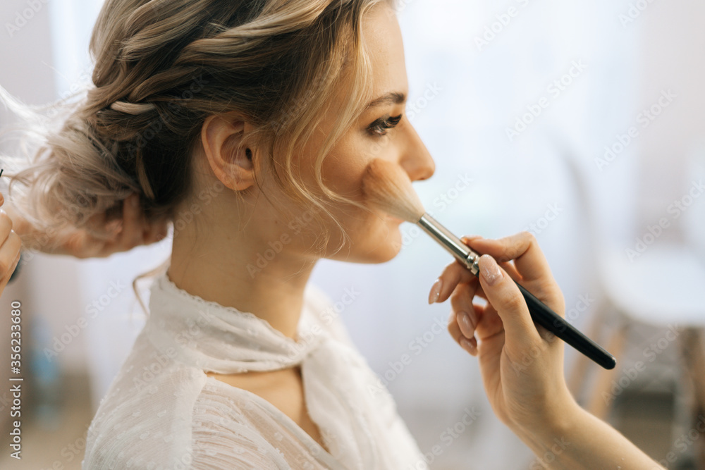 Fototapeta premium Close-up of make-up artist putting rouge on woman's cheek using brush while hairdresser making hair-do for young woman in beauty salon. Concept of backstage work.