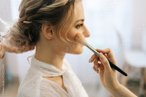 Close-up of make-up artist putting rouge on woman s cheek using brush while hairdresser making hair-do for young woman in beauty salon. Concept of backstage work.
