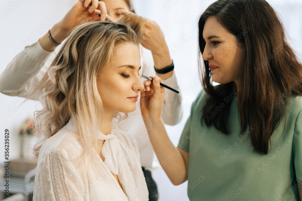 Professional make-up artist making face makeup while hairdresser making hair-do for young woman in beauty salon. Concept of backstage work. Professional make-up artist making face makeup while hairdresser making hair-do for young woman in beauty salon. Concept of backstage work.