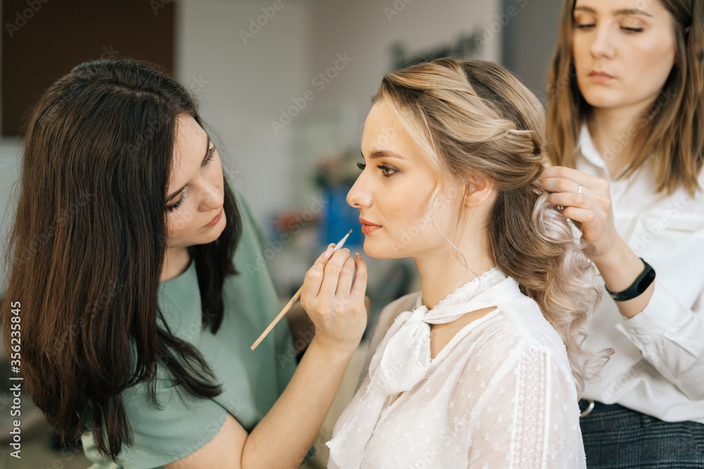 Make-up artist applying eyeshadows while hairdresser making hairstyle in dressing room in front of the mirror. Concept of backstage work. Make-up artist applying eyeshadows while hairdresser making hairstyle in dressing room in front of the mirror. Concept of backstage work.