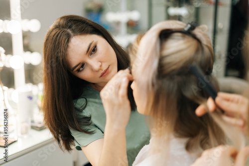 Close-up of preparation process of gorgeous young woman, hairdresser is twisting long fair hair, female make-up artist working in dressing room. Concept of backstage work.