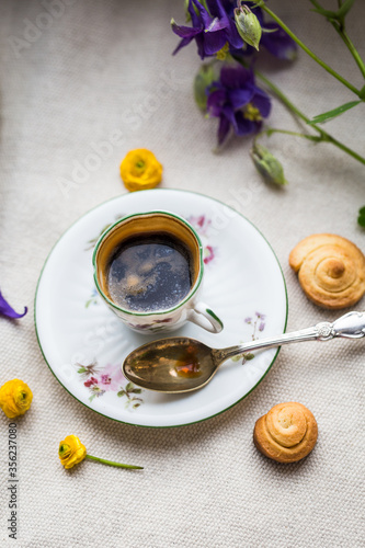 Espresso coffee in small delicate vintage cup with floral print, spoon, homemade cookies and spring flowers on linen background.