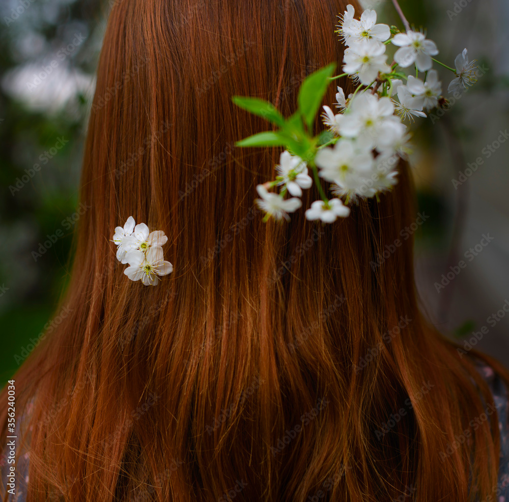 Naklejka premium a red-haired girl stands from back in a blooming garden with flowers in her hair