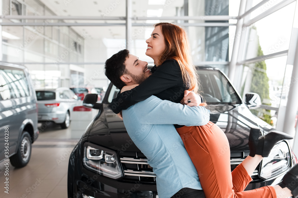 Guy hugs his girlfriend in a car dealership on the background of a purchased car Guy hugs his girlfriend in a car dealership on the background of a purchased car