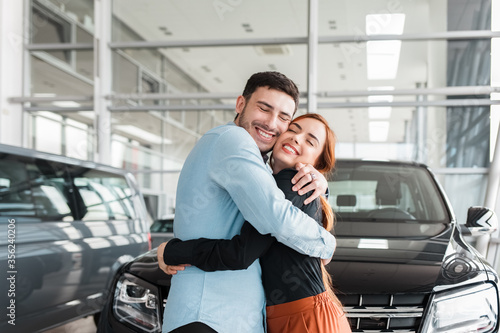 Guy hugs his girlfriend in a car dealership on the background of a purchased car