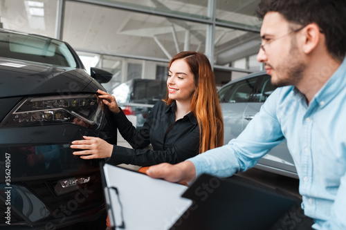 Salesman shows the car to a young client of a car dealership. Girl chooses a new car