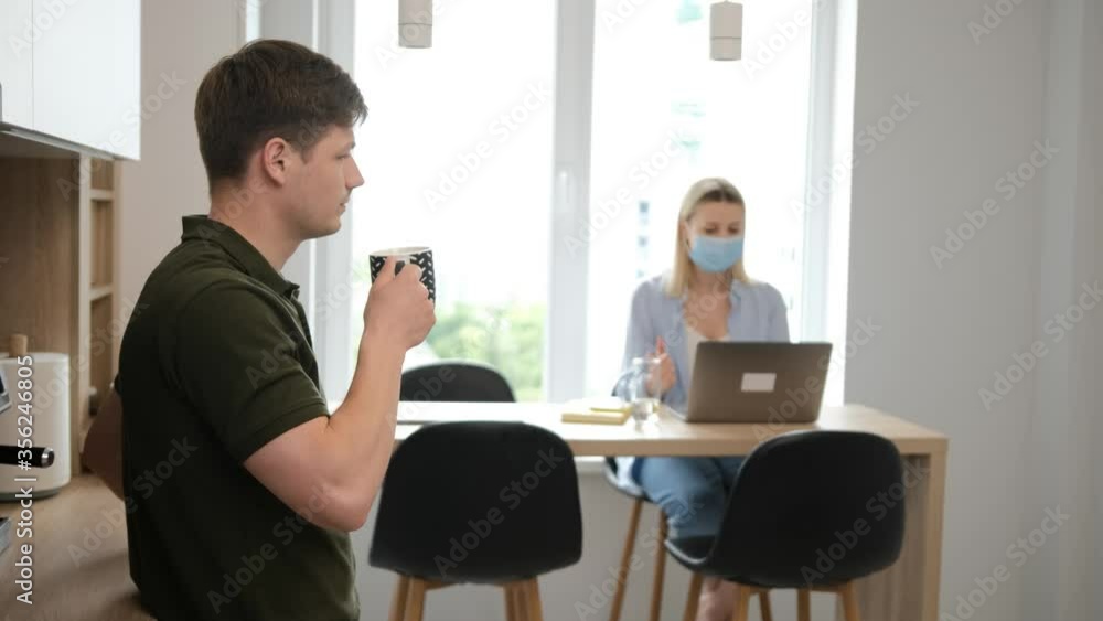 Man is drinking a cup of tea in the kitchen while his wife is working at the laptop with a medicinal mask on face. Stay at home and work from home concept Man is drinking a cup of tea in the kitchen while his wife is working at the laptop with a medicinal mask on face. Stay at home and work from home concept