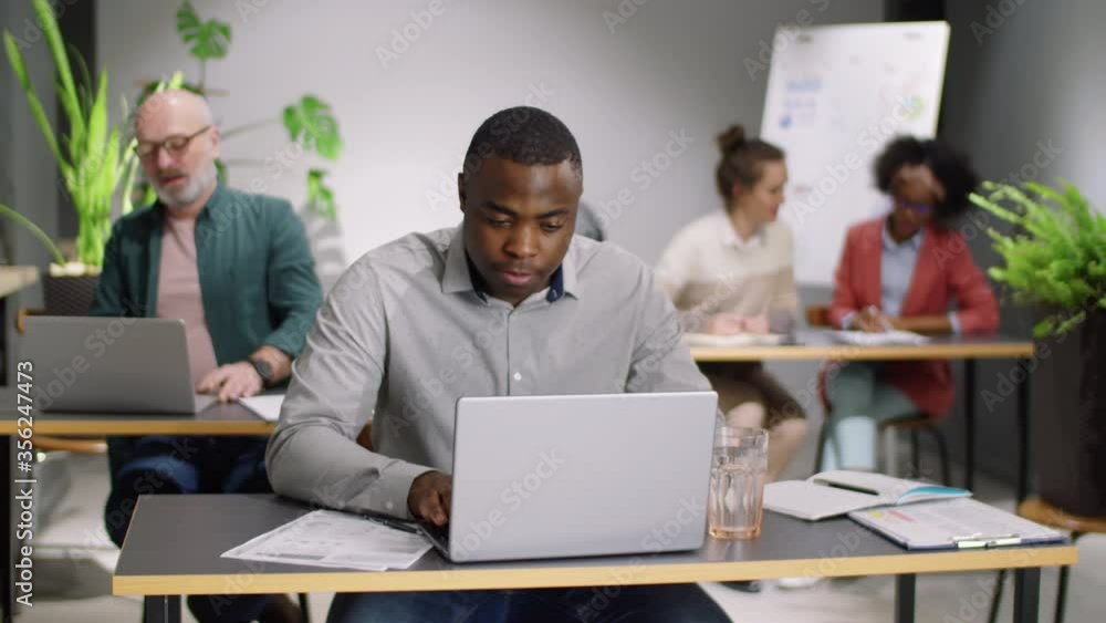 Young cheerful African American man working on laptop at desk, then looking at camera and smiling during workday in open space office with multiethnic mixed-age employees Young cheerful African American man working on laptop at desk, then looking at camera and smiling during workday in open space office with multiethnic mixed-age employees
