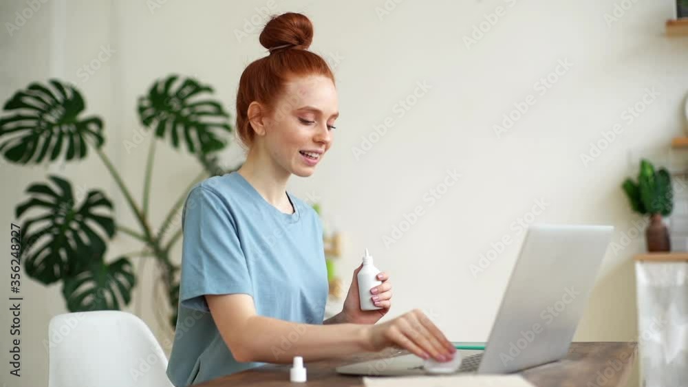Happy redhead young woman is cleaning keyboard of laptop computer with sanitizer before starting work in light room at the home office. Concept of remote work or education from home in self isolation Happy redhead young woman is cleaning keyboard of laptop computer with sanitizer before starting work in light room at the home office. Concept of remote work or education from home in self isolation