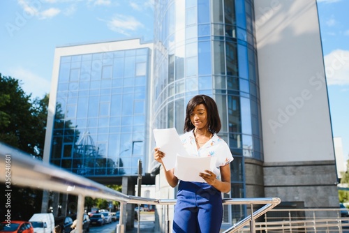 Portrait of young female African American job seeker keeping a folder with CV in her hands standing against office building. Blurred background with copy space