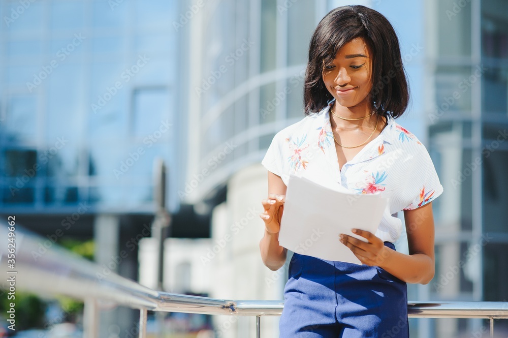 Portrait of young female African American job seeker keeping a folder with CV in her hands standing against office building. Blurred background with copy space