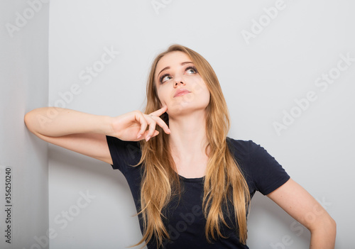 Confusion thinking with fair blond hair woman looking up with sad eyes and scratching the head. Casual neutral shirt clothing on grey studio wall for isolation portrait.