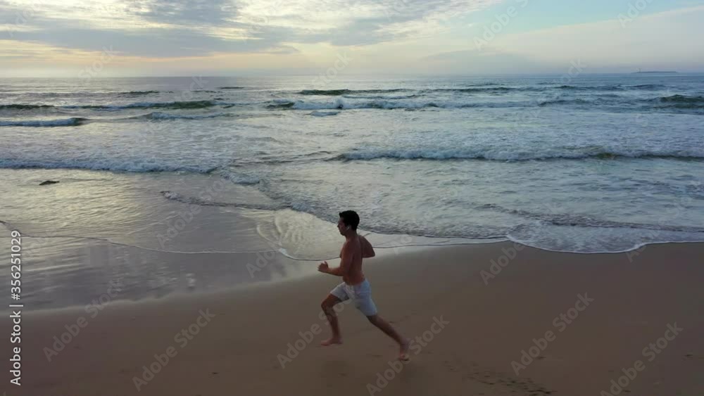 Aerial: Drone panning by shirtless man running on shore at beach during sunset - Punta del Este, Uruguay Aerial: Drone panning by shirtless man running on shore at beach during sunset - Punta del Este, Uruguay