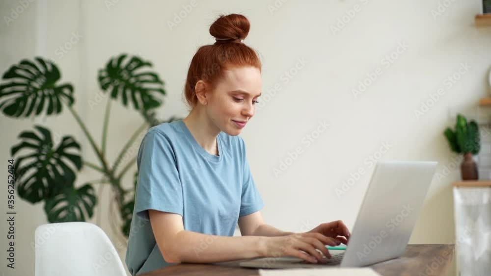 Cute young redhead woman is working on modern laptop at the desk in light cozy room at the home office. Female gesticulates approvingly after receiving an online answer. Shotting in slow motion. Cute young redhead woman is working on modern laptop at the desk in light cozy room at the home office. Female gesticulates approvingly after receiving an online answer. Shotting in slow motion.