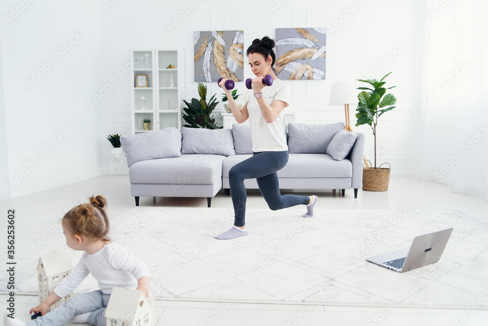 Sportive mother making exercises with dumbbells and her cute tender daughter playing with toys on foreground. Sportive mother making exercises with dumbbells and her cute tender daughter playing with toys on foreground.