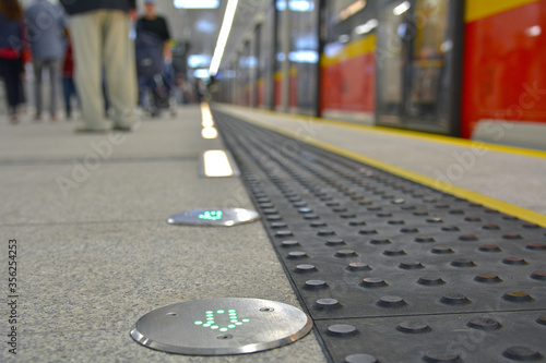 Concrete tactile strips with an yellow line - for visually impaired (handicapped) and people with blindness - on an station, with passengers and a subway train in the background