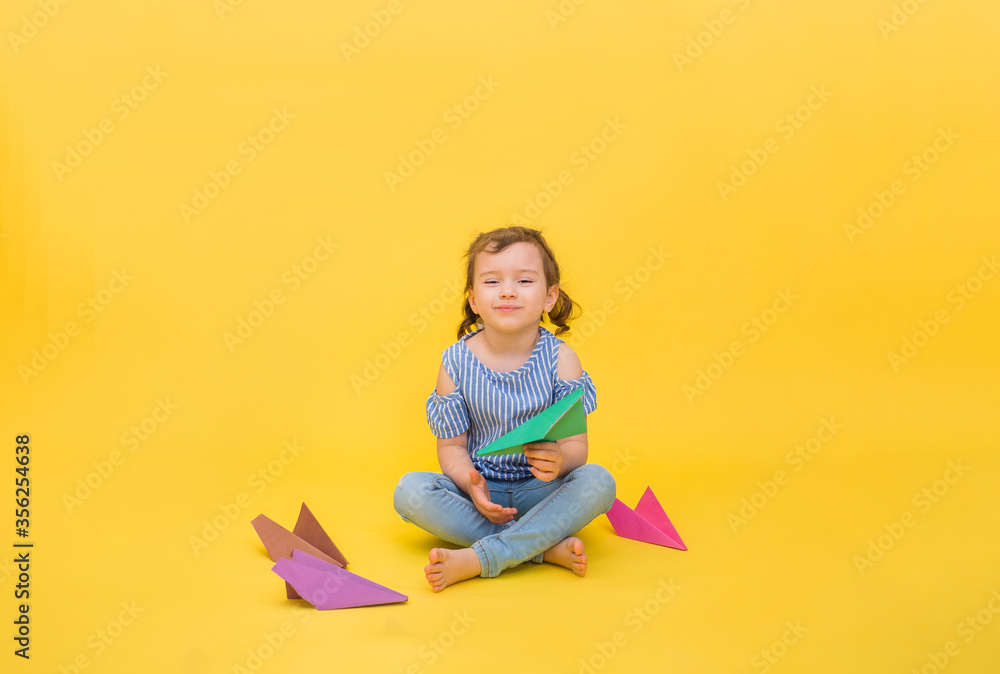 A happy girl sits with origami paper planes on a yellow background and laughs