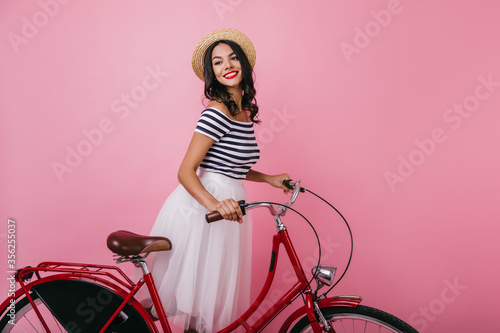 Fototapeta Naklejka Na Ścianę i Meble -  Inspired shapely woman standing on pink background with bicycle and looking away. Blissful brown-haired girl in hat enjoying indoor photoshoot.