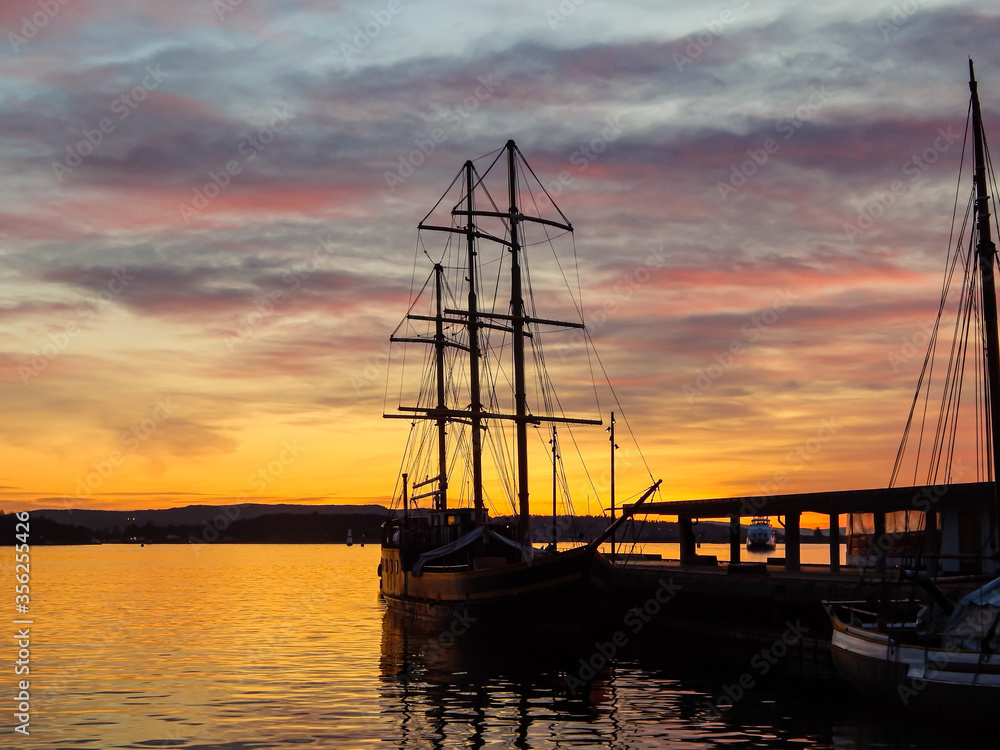 Dark ship in evening harbour. Beautiful sky with red clouds. Golden water reflections. Oslo, Norway.