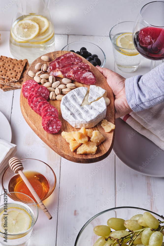 Girl prepares a romantic dinner for two. Cheese and sausage delicacies, nuts, fruits, olives, bread, grapes, wine and honey on an old white wooden table.