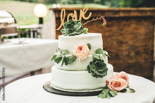 Close up of white wedding cake laying on table. Love inscription, pink rose and green leaves floral decoration. Outside terrace. Wedding day concept.