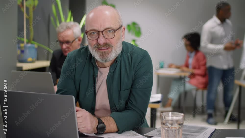 Senior Caucasian man in glasses taking notes and then looking at camera and smiling while working at desk in open space office with diverse colleagues Senior Caucasian man in glasses taking notes and then looking at camera and smiling while working at desk in open space office with diverse colleagues