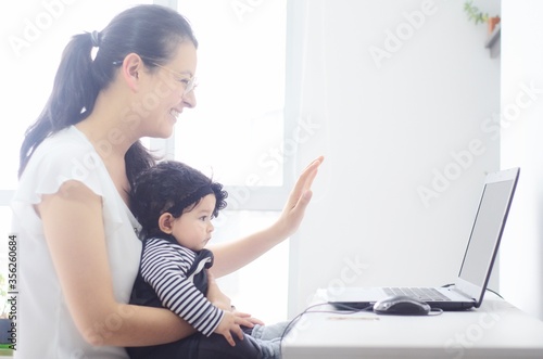 Feliz madre y su bebé usan la computadora para hacer video llamada desde su casa. Madre y su hija saludando con la mano a su familia mediante una video llamada. Uso de tecnologías para la comunicación