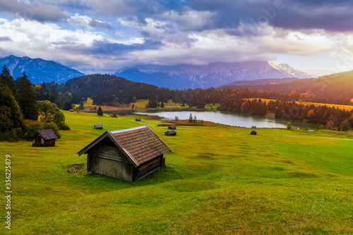 Panoramic morning scene of Wagenbruchsee (Geroldsee) lake with Zugspitze mountain range on background. Amazing autumn view of Bavarian Alps, Germany, Europe. Wooden hut on meadow by Geroldsee lake.
