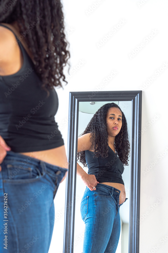 Beautiful African American woman and mother stands in front of a mirror and  attempts to try to close her tight fitting blue jeans pants after postpartum  diet and weight loss exercise. Stock