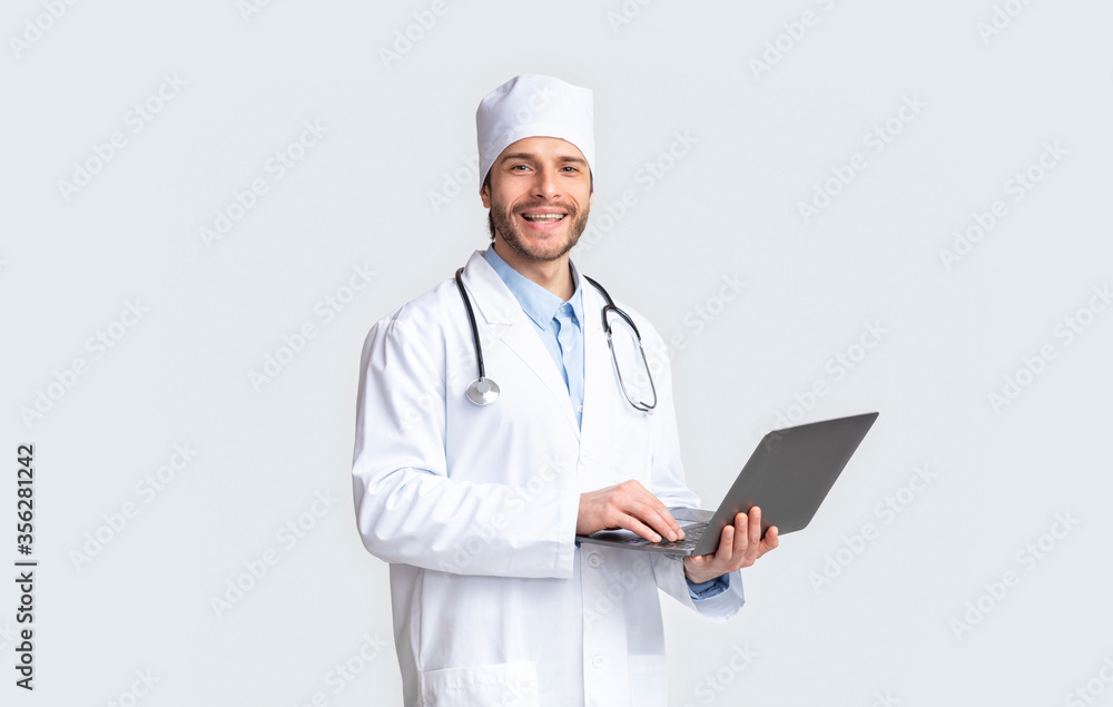 Friendly doctor smiling with laptop, grey studio background