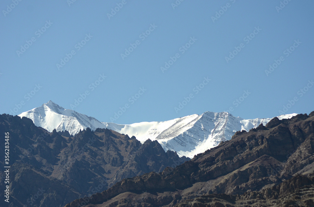 Mountain peaks of the Western Himalayas are seen against a clear blue sky. They contrast with the dark rocky mountains in the foreground.