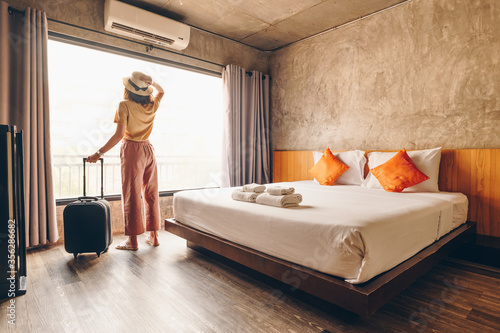 Portrait of tourist woman standing nearly window, looking to beautiful view with her luggage in hotel bedroom after check-in. Conceptual of woman lifestyle when traveling on her vacation.