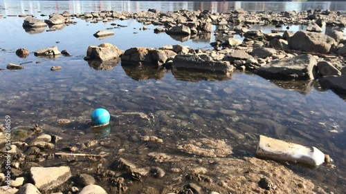 Rocky riverbank. Polluted shore with foam on the water. Dirty plastic bottles and a blue ball float in the water