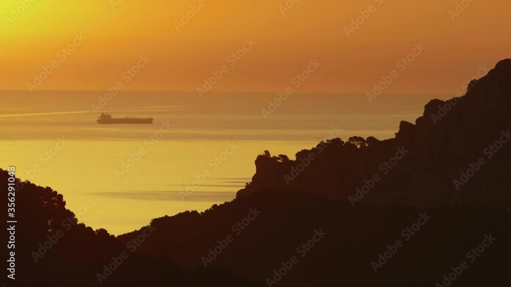 An oil tanker in the distance under an orange sky viewed through hillsides.