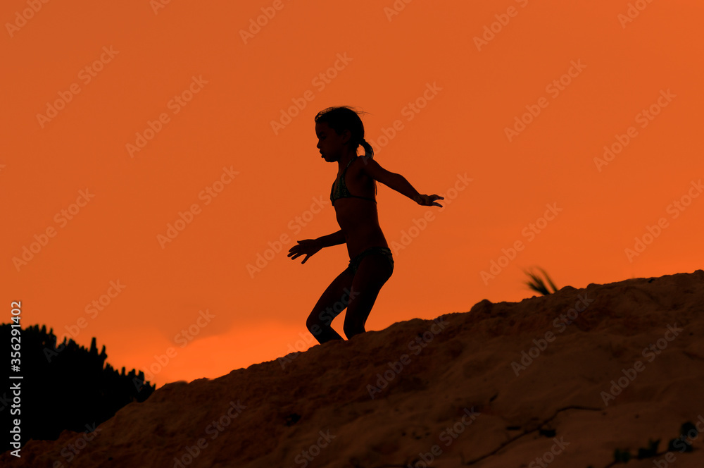 Silueta de niña corriendo sobre duna de playa en atardecer naranja durante vacaciones de verano.