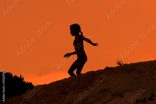 Silueta de niña corriendo sobre duna de playa en atardecer naranja durante vacaciones de verano. © Iván Gabaldón