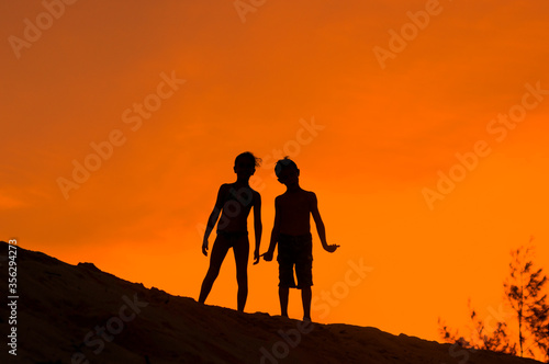Siluetas de niña y niño de pie sobre duna de playa en atardecer naranja, vacaciones de verano.