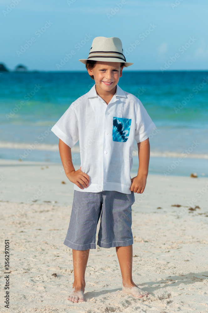 Niño con pantalones cortos y camisa blanca con sombrero fedora en playa del mar Caribe durante vacaciones de verano.